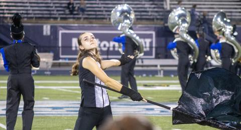 Marching band sousaphone players face backfield while a colorguard soloist spins a black flag