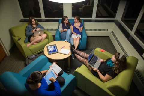 A group of students sit in chairs talking, reading and working on computers