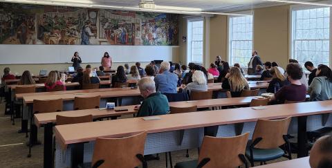 An audience in a lecture hall listens to two female speakers at the front of the room