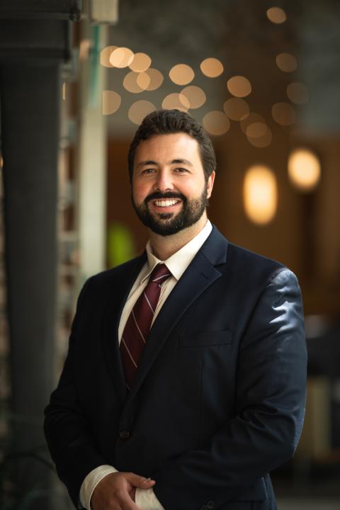 EdD student Cayce smiling directly at camera in front of a blurry background, wearing a navy suit and red tie. 