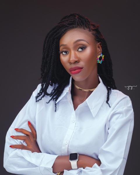 PhD student Mariama Darboe standing in front of plain background, wearing a white button down shirt, crossing her arms and looking at the camera