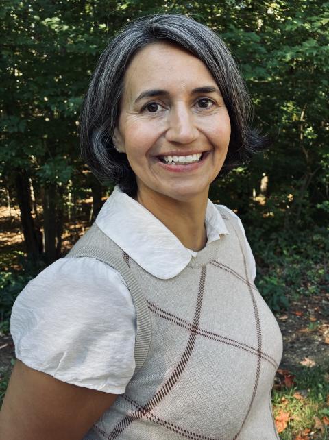 EdD student Melissa Gianino stands, smiling in front of a forest background, wearing a grey sweater vest and collared white shirt